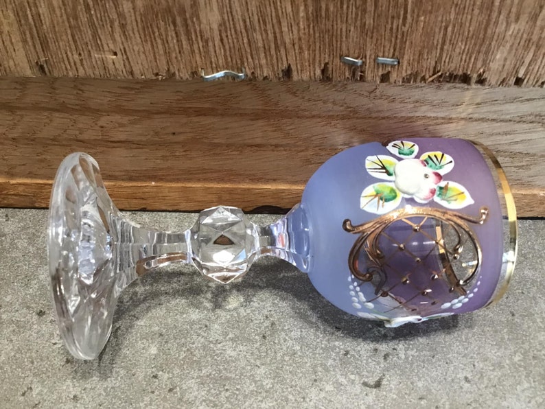 May include: A purple glass goblet with a gold rim and hand-painted floral design. The goblet has a clear glass stem and base.