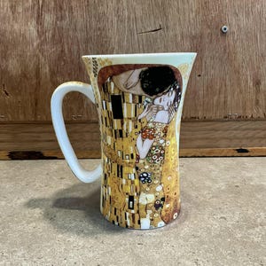 May include: A white ceramic mug with a flared design features a colourful reproduction of Gustav Klimt's "The Kiss" painting.