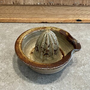 May include: A ceramic citrus juicer with a light beige interior and a dark brown rim. The juicer has a conical reamer in the center and a spout for pouring. The stoneware piece is handmade and has a rustic appearance.