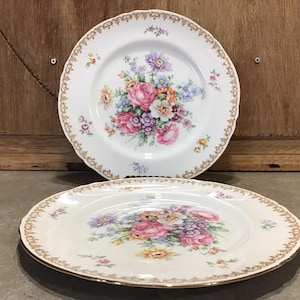 May include: Two white ceramic plates with gold trim and a floral design. The plates feature a bouquet of pink, yellow, and purple flowers.