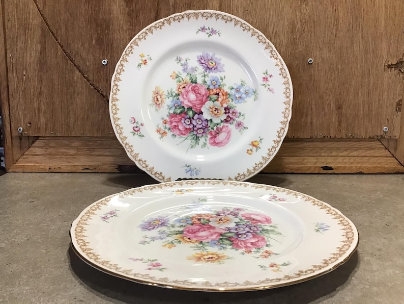 May include: Two white ceramic plates with gold trim and a floral design. The plates feature a bouquet of pink, yellow, and purple flowers.