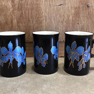 May include: Three black ceramic mugs with blue and gold orchid designs. The mugs have white interiors and are arranged in a row on a light brown surface.