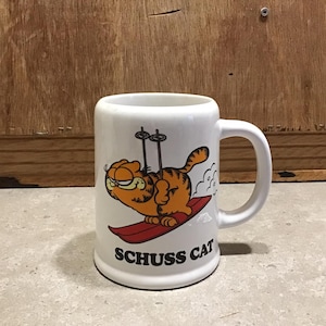 May include: White ceramic mug with a cartoon image of Garfield the cat skiing. The mug has the text "Schuss Cat" printed on the bottom.