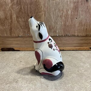 Könnte beinhalten: Eine weiße Keramik-Hundefigur mit schwarzen und rosafarbenen Akzenten. Der Hund sitzt mit dem Kopf nach oben geneigt und leicht geöffnetem Maul. Die Figur ist mit einem floralen Muster an der Seite bemalt.