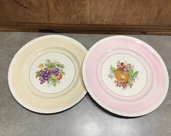 Puede incluir: Dos platos de cerámica vintage con fondo blanco y borde decorativo. Un plato tiene un borde rosa y presenta una imagen pintada de fruta, incluyendo una naranja, uvas y bayas. El otro plato tiene un borde crema y presenta una imagen pintada de uvas y una pera.