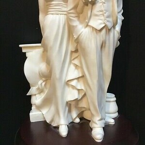 May include: A white porcelain figurine of a couple standing together. The woman is wearing a long dress with a ruffled skirt and the man is wearing a suit with a tie. The figurine is on a wooden base.
