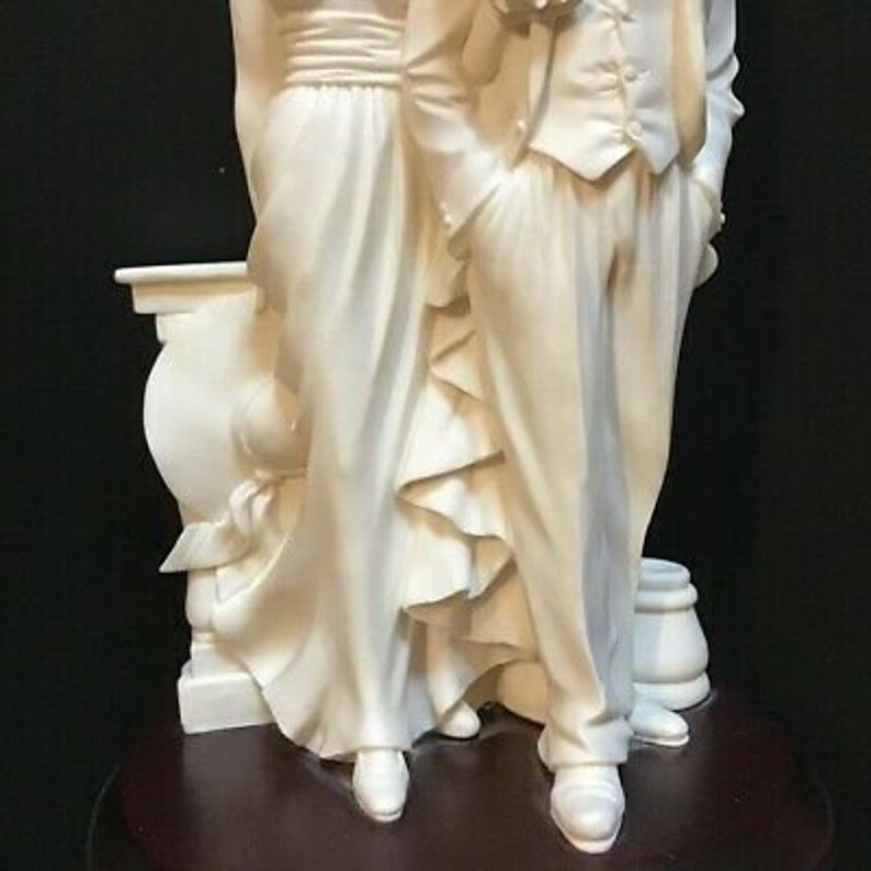 May include: A white porcelain figurine of a couple standing together. The woman is wearing a long dress with a ruffled skirt and the man is wearing a suit with a tie. The figurine is on a wooden base.
