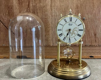Op de afbeelding: Een gouden klok met een glazen stolp, met een witte wijzerplaat met zwarte Romeinse cijfers en zwarte wijzers. De klok heeft een klein bloemenmotief en de tekst "Quartz" op de wijzerplaat. De klok staat op een gouden voet met een decoratieve bol.