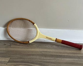 以下が含まれることがあります： 木製のフレームとクリーム色のハンドルが付いたヴィンテージテニスラケット。ラケットは赤いグリップと白いストリングスを備えています。フレームはライトブラウンで、赤いアクセントが付いています。ラケットは壁に立てかけられています。
