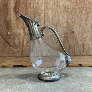 Könnte beinhalten: Eine klare Glaskaraffe mit einem silberfarbenen Metalldeckel und Griff. Die Karaffe ist vogelförmig und hat einen runden Boden.