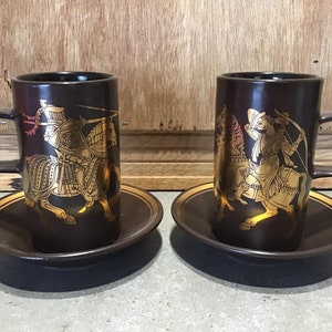 May include: Two brown ceramic coffee mugs with gold trim and a gold design of a knight and a woman on horseback. Each mug is on a matching saucer.