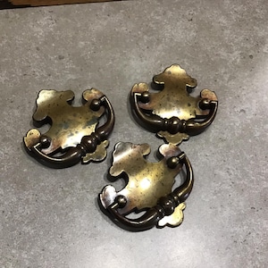 May include: Three antique brass cabinet pulls with ornate designs. Each pull has a curved handle and a decorative backplate.