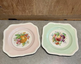 Puede incluir: Dos platos decorativos con bordes festoneados. Uno es rosa y el otro verde claro. Cada plato presenta un diseño central de frutas con una naranja, uvas y otras frutas. Los platos tienen un borde blanco en forma de cuerda.