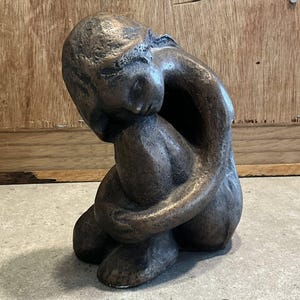Op de afbeelding: Bronzen sculptuur van een persoon in een zittende, foetale positie. Het hoofd van de figuur rust op de knieën, met armen om de benen. De sculptuur heeft een getextureerd oppervlak en staat tegen een neutrale achtergrond.