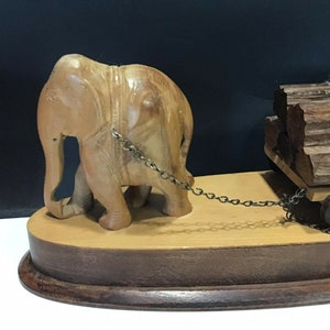 May include: A wooden elephant figurine pulling a wooden cart loaded with logs. The elephant is carved in detail and has a chain attached to the cart. The cart has a wooden wheel and is on a wooden base.