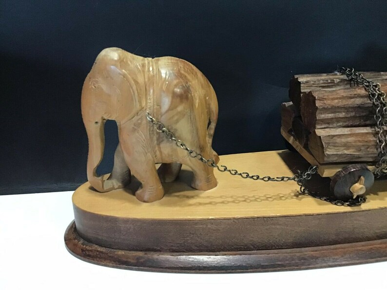 May include: A wooden elephant figurine pulling a wooden cart loaded with logs. The elephant is carved in detail and has a chain attached to the cart. The cart has a wooden wheel and is on a wooden base.