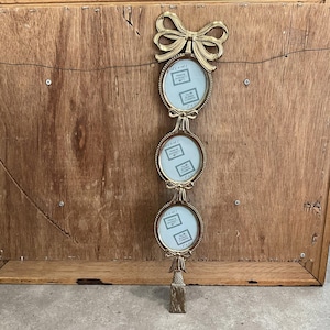 May include: A gold-coloured triple photo frame with oval frames and bow details. The frames are linked by decorative bows and a tassel at the base. Each frame holds a small photo, with the text "2 x 3" visible.