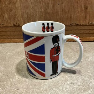 Puede incluir: Taza de cerámica blanca con un diseño de la bandera británica y una ilustración de un guardia real con uniforme rojo. La taza tiene un asa blanca y un borde dorado. La letra "N" es visible en la taza.