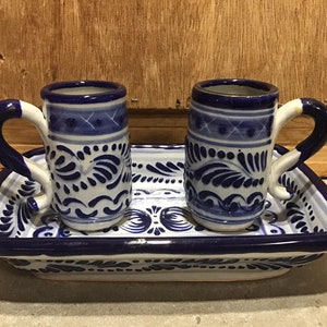 May include: A set of two small, blue and white ceramic mugs with curved handles, resting on a matching rectangular tray. The mugs and tray feature a detailed floral and geometric pattern. The set is designed with a traditional aesthetic.