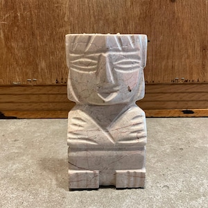 May include: A light pink and beige stone sculpture carved into the shape of a stylized human figure with a rectangular head, almond-shaped eyes, a prominent nose, and arms crossed over the chest, featuring visible natural veining throughout.