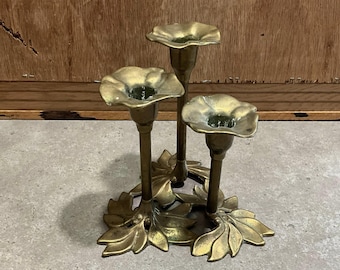 Könnte beinhalten: Ein Messing-Kerzenhalter mit drei blütenförmigen Kerzenschalen. Der Kerzenhalter hat einen Sockel mit Blattdetails. Das Messing hat einen goldenen Farbton. Der Kerzenhalter ist für drei Kerzen konzipiert.