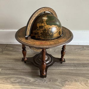 May include: An antique globe on a wooden stand. The globe features a detailed map with brown and green tones. The stand has a circular base and four legs, all in a dark brown finish. A curved, labeled band encircles the globe.