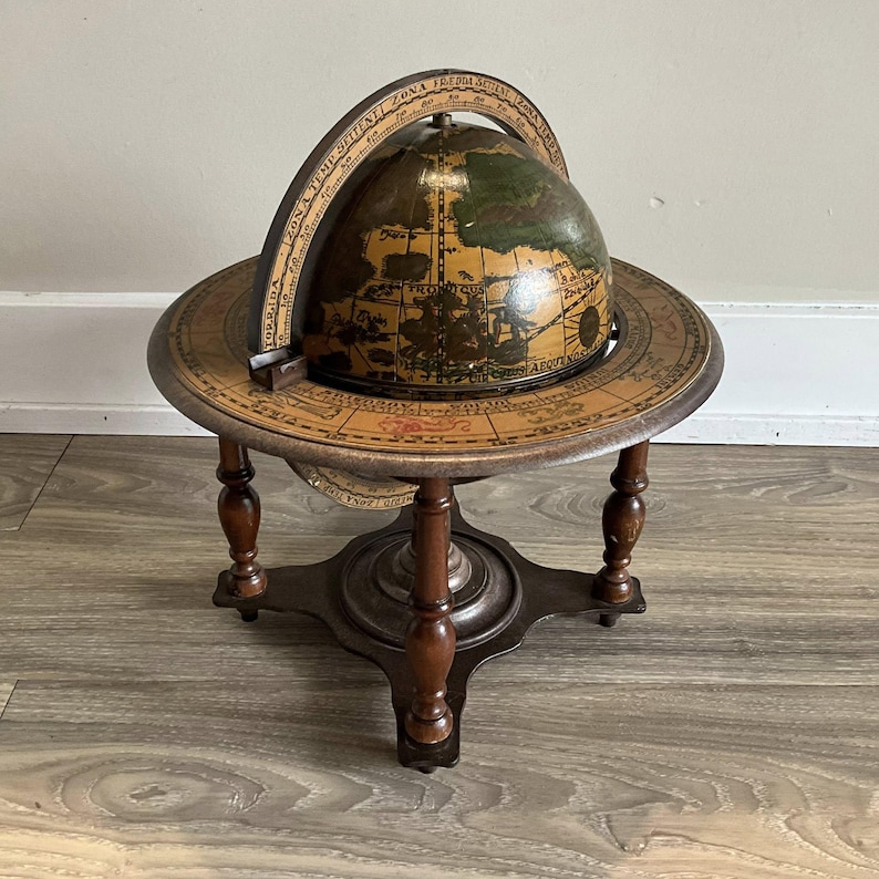 May include: An antique globe on a wooden stand. The globe features a detailed map with brown and green tones. The stand has a circular base and four legs, all in a dark brown finish. A curved, labeled band encircles the globe.