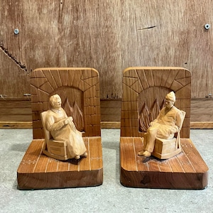 May include: A pair of wooden bookends, each with a carved design. One shows a person in a rocking chair, the other a person in a chair, both in front of a fireplace. The bookends are made of light brown wood.