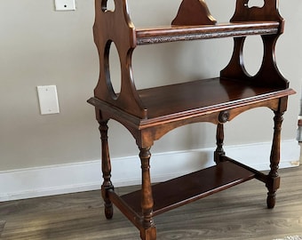 May include: A dark brown wooden two-tiered shelf with decorative cutouts and turned legs. The shelf has a carved design on the top edge.