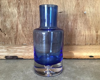 Könnte beinhalten: Eine kobaltblaue Glasvase mit rundem Korpus und einem klaren Glasfuß. Die Vase hat einen kleinen, klaren Glasknopf am Boden.