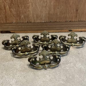 以下が含まれることがあります： 装飾的で曲線的なデザインの、真鍮製のアンティーク引き出しプルが6つ。各プルは中央にハンドルと装飾的な装飾が施されています。真鍮は暖かく、古びた風合いで、ヴィンテージスタイルを連想させます。これらのプルは、家具の修復や装飾プロジェクトに使用されます。