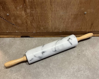 May include: A white marble rolling pin with a wooden handle. The rolling pin is lying on a gray surface.