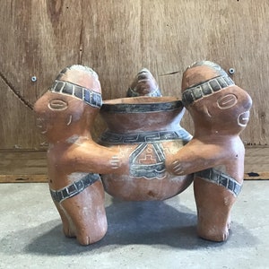 Könnte beinhalten: Eine Terrakotta-Keramikskulptur von zwei Figuren, die einen großen Topf tragen. Die Figuren sind mit geometrischen Mustern gestaltet und der Topf hat ein geometrisches Muster.