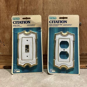 May include: Two Hall Mack Citation switch plates. One is a switch plate and the other is a duplex receptacle plate. Both are white with gold trim and are on a blue and white striped background.