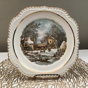 May include: A white ceramic plate with a gold rim features a winter scene with a farmhouse and snow-covered trees. The plate has the text "The Farmer's Home - Winter" printed on the bottom.