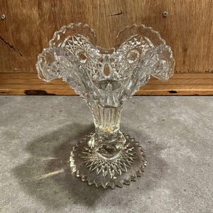 May include: Clear glass pedestal vase with a ruffled rim and intricate cut glass patterns. The vase has a wide, starburst base and a detailed stem. The design features diamond and fan-shaped cuts, creating a vintage aesthetic. Ideal for displaying flowers or as a decorative piece.