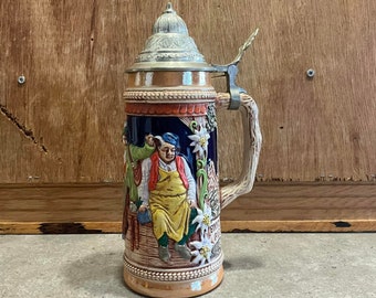 May include: A ceramic beer stein with a pewter lid and handle. The stein features a colorful scene of two men in traditional clothing, with a mountain and flowers in the background. The text "Servus" is visible on the stein.