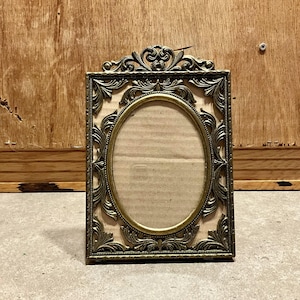 May include: Ornate, antique-style picture frame with an oval opening. The frame is made of a gold-colored metal with intricate floral and scrollwork designs. The background is a light tan color, and the frame is set against a wooden surface.