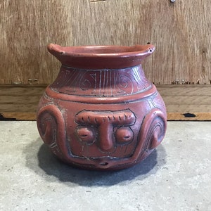 以下が含まれることがあります： 正面に顔が彫られた、小さな赤い粘土の壺。壺は口が広く、丸い胴体をしている。顔には目、鼻、口がある。