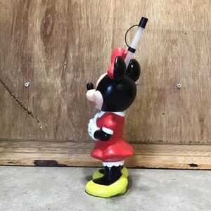 May include: A Minnie Mouse-shaped tumbler with a red dress, black ears, and a red bow. The tumbler has a clear straw and a yellow base. The character is standing on a yellow base. The tumbler is made of plastic.