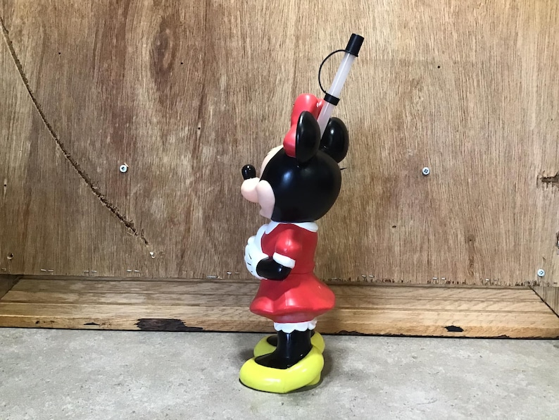 May include: A Minnie Mouse-shaped tumbler with a red dress, black ears, and a red bow. The tumbler has a clear straw and a yellow base. The character is standing on a yellow base. The tumbler is made of plastic.