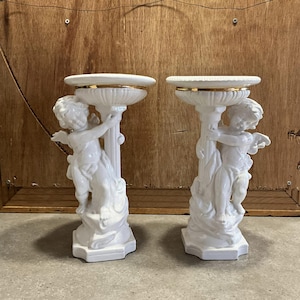 Könnte beinhalten: Ein Paar weiße Keramiksockel mit Cherubfiguren und goldenen Verzierungen. Jeder Sockel hat eine runde Oberseite und eine Basis mit einer Cherubfigur, die den Sockel festhält.