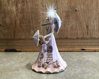 May include: A porcelain figurine of two women holding parasols. The women are wearing pink dresses with floral patterns. The larger woman is holding a parasol with a white feather on top. The smaller woman is holding a parasol with a white trim. The base of the figurine is decorated with flowers and the text "Daughters Blossom in the Garden of..."