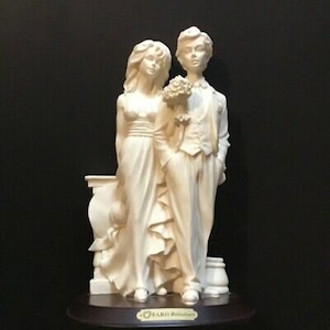 May include: A white porcelain figurine of a bride and groom standing together. The bride is wearing a flowing gown and the groom is wearing a tuxedo. They are holding a bouquet of flowers. The figurine is on a wooden base.