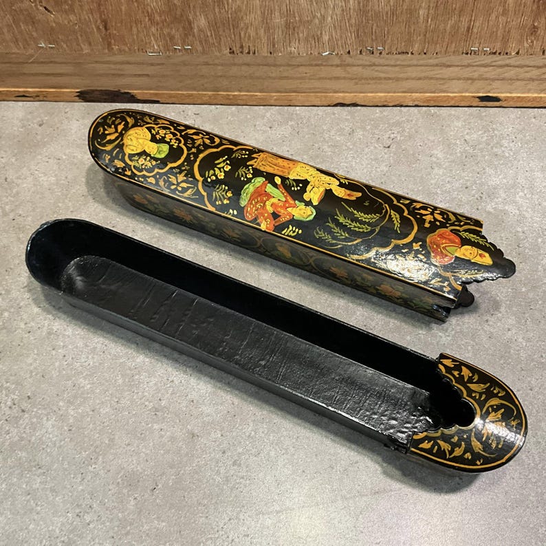 May include: A black, oblong, decorative box with a lid, featuring intricate gold and colorful painted designs. The lid is open, revealing the interior. The box is likely used for storage or display.