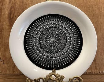 May include: A white ceramic plate featuring a black and white mandala design. The plate is displayed on a gold metal stand.