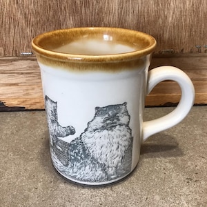 Puede incluir: Taza de cerámica blanca con un borde marrón y un diseño de tres gatos. Los gatos están dibujados en blanco y negro y están en varias poses.