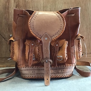 Puede incluir: Una mochila de cuero marrón con intrincados detalles tejidos y una solapa con cierre de hebilla. La mochila tiene dos bolsillos grandes en la parte delantera y un cierre de cordón en la parte superior.