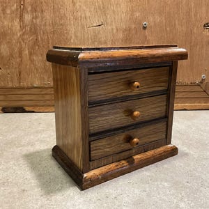 May include: A small, wooden jewelry box with three drawers. The box is made of dark oak wood with a natural finish. Each drawer has a small, round wooden knob. The box has a rectangular shape and is designed to sit on a flat surface.