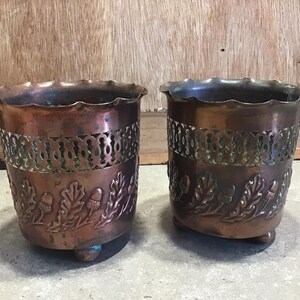 May include: Two copper plant pots with an acorn and leaf design. The pots have a scalloped rim and stand on three small feet.